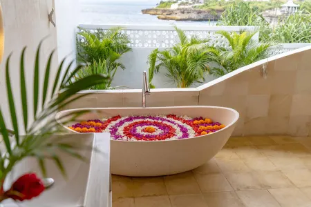 Luxurious outdoor bathtub filled with floating colorful flower petals, overlooking tropical ocean cliffs and lush greenery. - Bali Villas